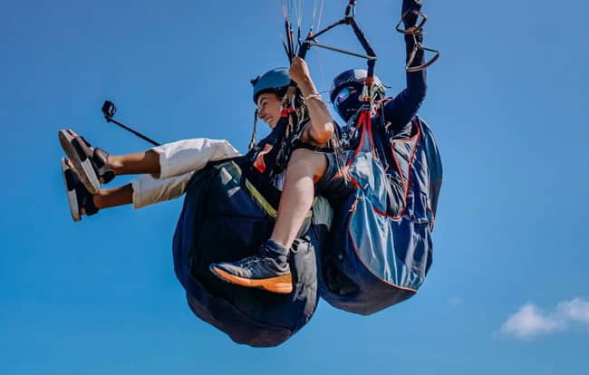 Deux personnes en parapente avec un bâton à selfie contre un ciel bleu clair.