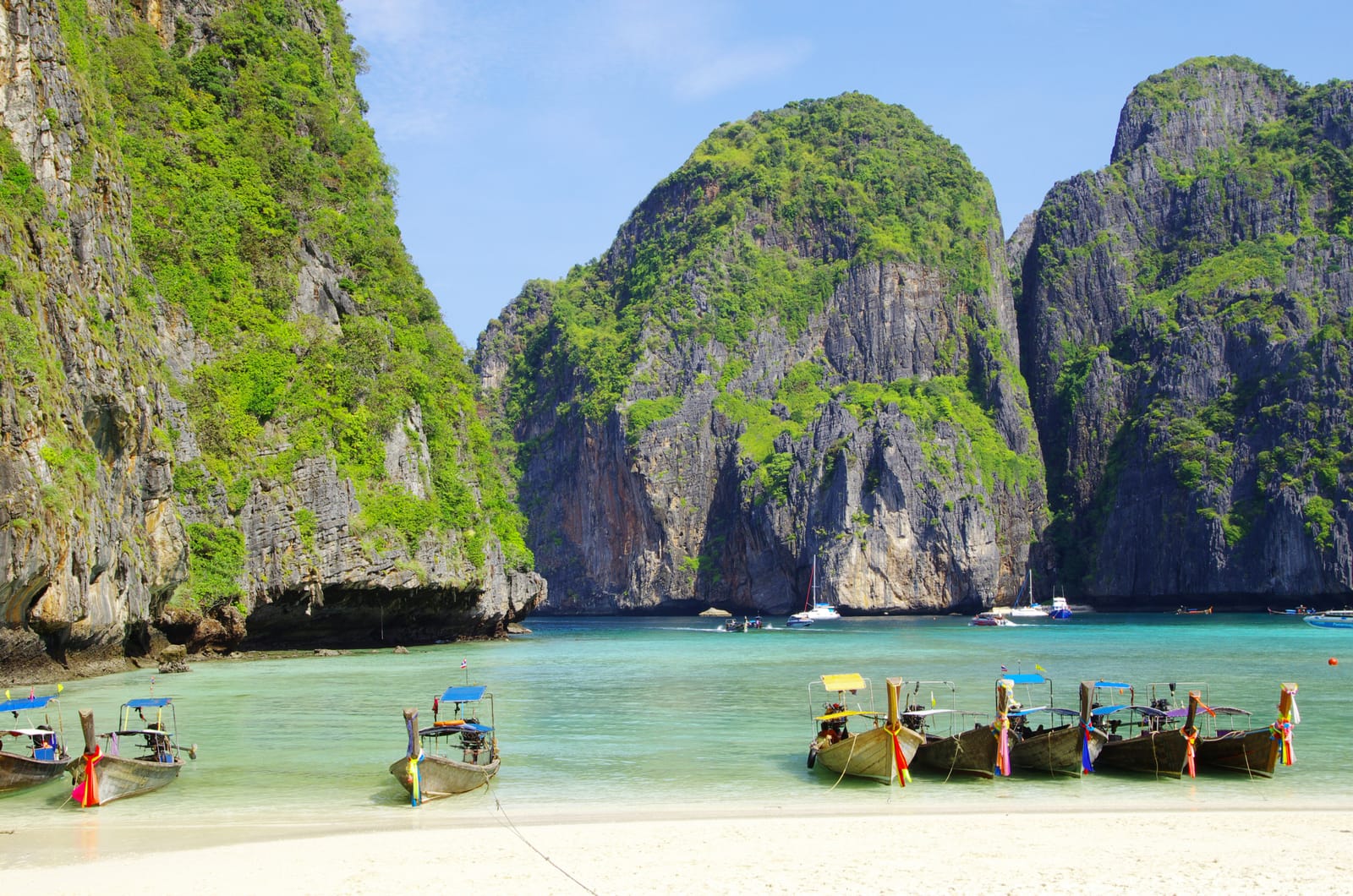 Des bateaux traditionnels en bois bordent une plage de sable aux eaux turquoise claires, entourée de falaises calcaires abruptes et couvertes de verdure sous un ciel bleu.