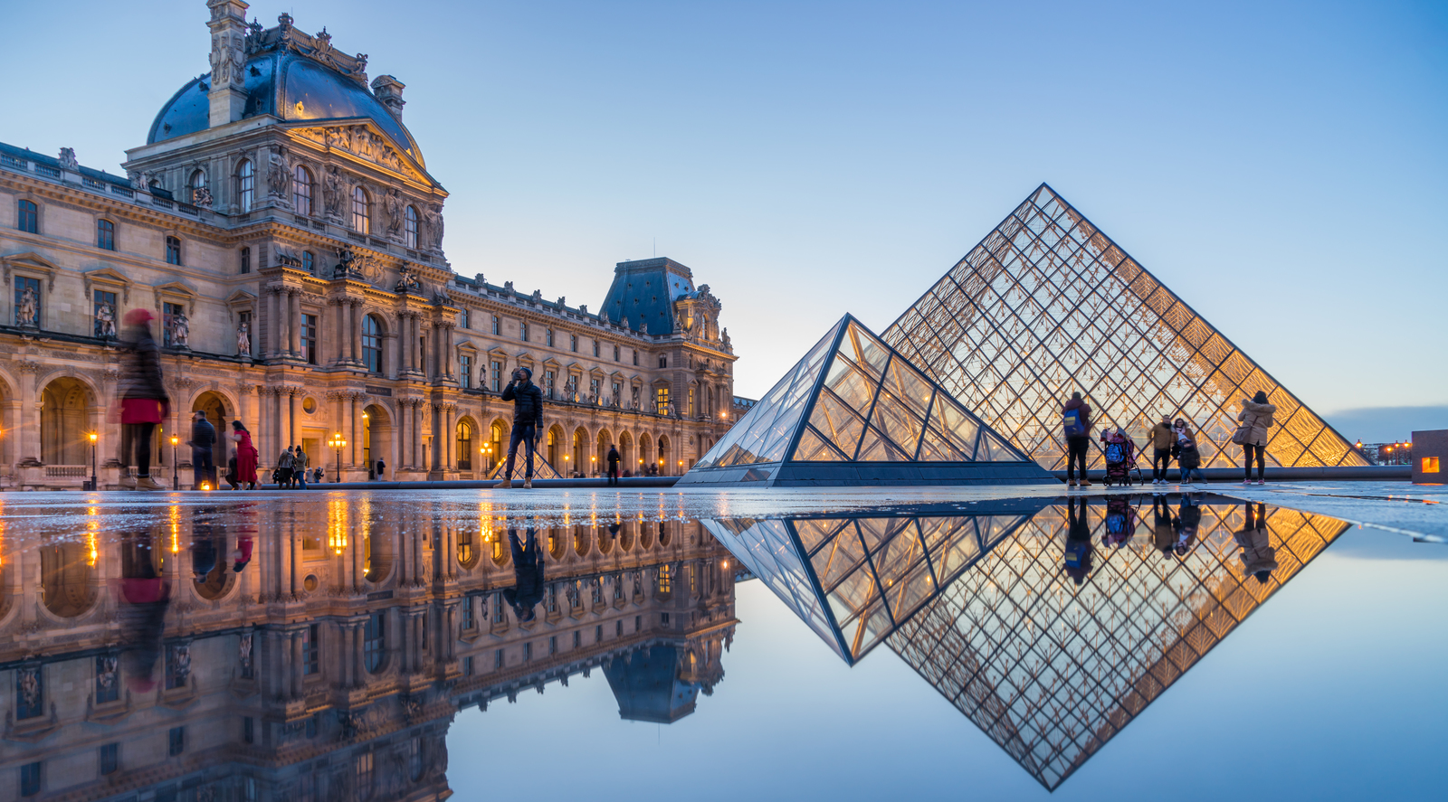 Les gens marchent près de l'entrée de la pyramide de verre du musée du Louvre à Paris au crépuscule, avec le bâtiment et la pyramide reflétés dans une grande flaque d'eau.